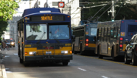 A Seattle Metro bus travels down a city street, with cyclists and cars nearby. The front of the bus displays a sign reading "Seattle." Other buses are visible in the background.