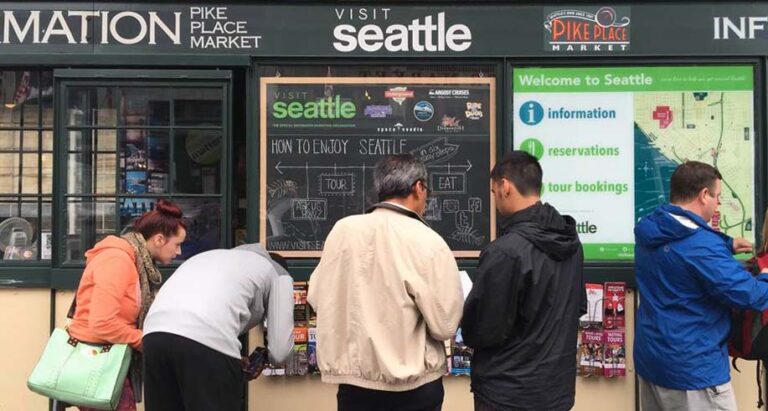 People standing in front of a tourist information booth in Seattle, checking maps and brochures. The booth displays signs for Pike Place Market and offers information, reservations, and tour bookings.