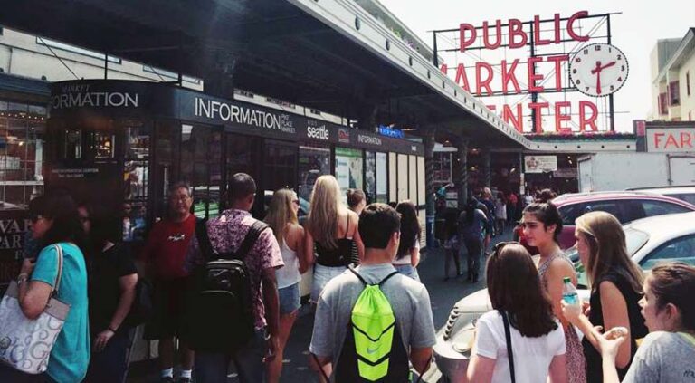 People gather outside the entrance of a bustling market, with a large sign reading "Public Market Center" and a clock. Stalls and informational booths are visible, and various people are walking or standing in the sunny outdoor area.