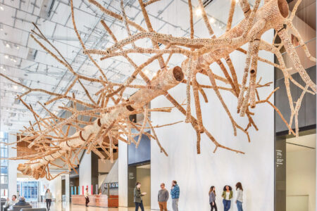 An intricate tree branch sculpture hangs from a high ceiling in a modern, spacious lobby. Five people walk beneath it on the polished floor. The sculpture is detailed, with numerous smaller branches extending outward, capturing attention.