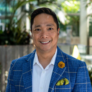 A person wearing a blue checkered blazer, white shirt, and yellow pocket square with a matching flower pin is smiling in an outdoor setting with greenery in the background.