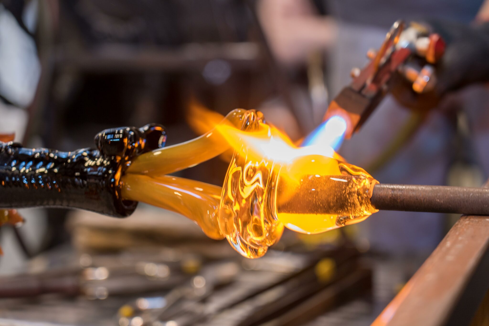 A glassblower shapes molten glass using a blowpipe and flame. The bright orange glass glows as it is heated and molded. Tools and equipment are visible in the background, suggesting a workshop setting.