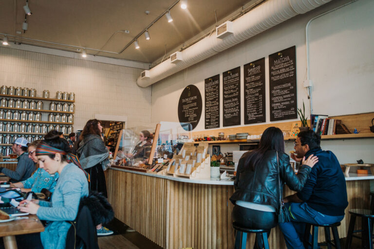 A cozy cafe scene with customers sitting at tables and the counter. A menu board is displayed above the counter. People are engaged in conversations and working on laptops. Shelves in the background hold jars and plants add to the decor.
