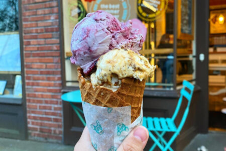 A hand holding a waffle cone with two scoops of ice cream, one purple and one beige with chocolate chunks. The background shows a brick storefront and a blue chair and table set on the sidewalk.