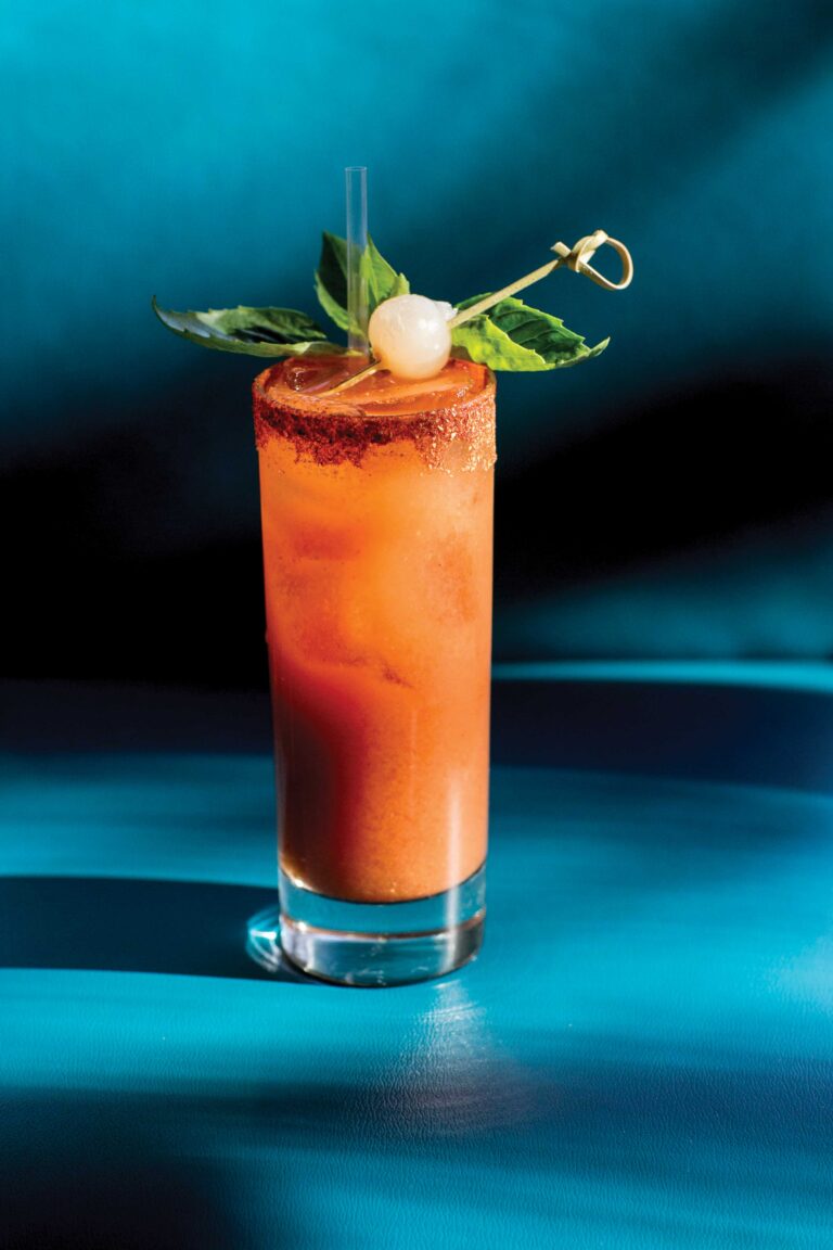 A tall glass filled with a vibrant orange cocktail, garnished with green leaves and a fruit skewer. The drink has a chili powder rim and is set against a rich blue background.