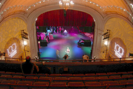 A theater interior with an ornate ceiling and chandelier. The stage is lit with colorful lights and a large red curtain. There are "1000 Doors" logos projected on the walls. A few people are near the stage, and the seats are mostly unoccupied.