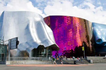 A modern, abstract building with a wavy, metallic exterior in silver and vibrant pink hues, set against a partly cloudy sky. People are walking in front of the building.
