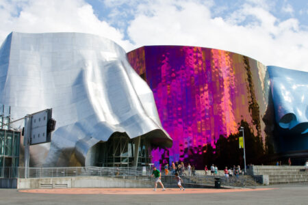 Colorful, abstractly shaped building with silver, pink, and blue metallic panels. People walk nearby on a sunny day. Cloudy sky in the background.