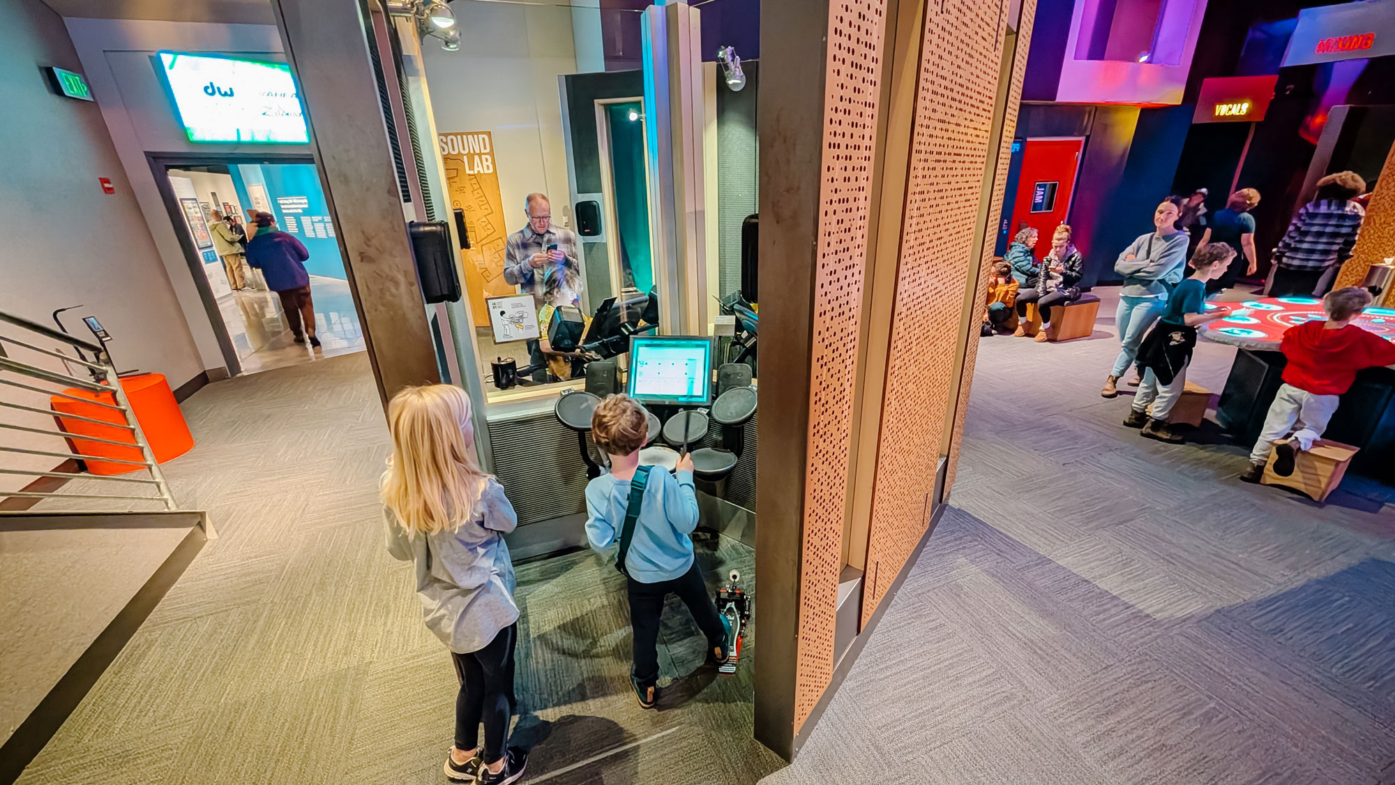 Children explore a hands-on science exhibit. A boy uses a virtual drum set, while others interact with displays in the background. The room features modern decor and various interactive stations.