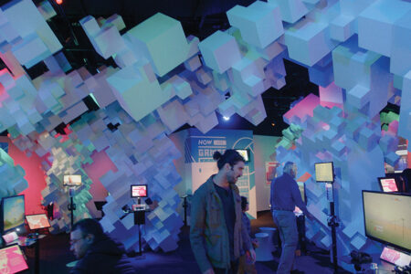 A modern exhibit featuring a ceiling of illuminated 3D cubes in blue hues. Visitors interact with digital screens and displays underneath the installation. The atmosphere is futuristic and engaging.