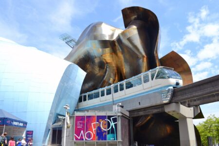 A white and blue monorail train passes through a futuristic, metallic building with curved surfaces under a bright blue sky with scattered clouds. People stand near colorful signage at the entrance.