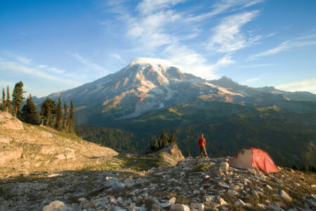 A person stands near a red tent, overlooking a rocky landscape with a towering snow-capped mountain under a clear blue sky. Tall pine trees are visible to the left, creating a serene camping scene.