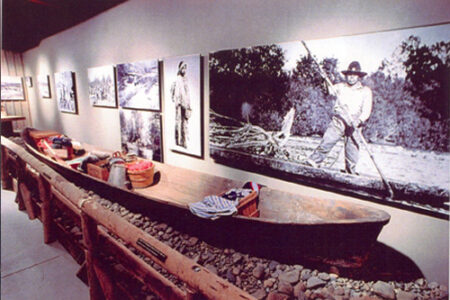 A museum exhibit features a long wooden canoe surrounded by various artifacts. Black-and-white photographs on the wall depict people in historical settings related to the canoe. The display is set against a dimly lit background.