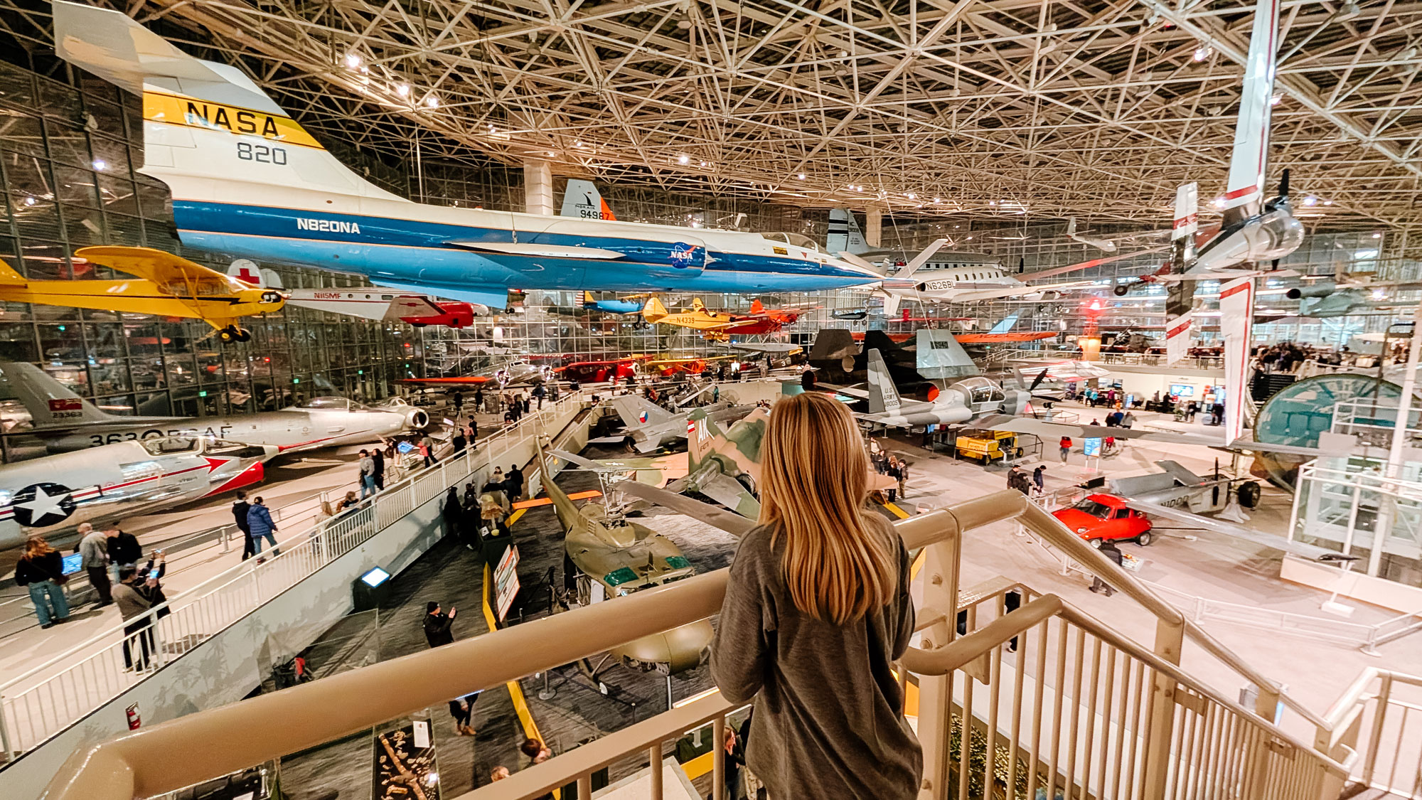 A person with long hair stands on a balcony inside a large museum, observing numerous airplanes displayed. The museum has high ceilings with a grid structure, and various aircraft, including a NASA plane, are showcased throughout the space.