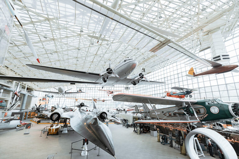 A large indoor aviation museum showcasing various aircraft on display, including a suspended airplane and several planes on the ground, all under a white, airy ceiling structure with large windows.