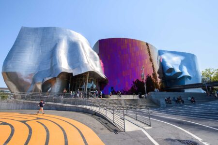 A vibrant, modern building with an abstract design featuring mirrored silver and iridescent panels. It sits under a clear blue sky with a few people walking and sitting nearby on the steps and ramps in the foreground.