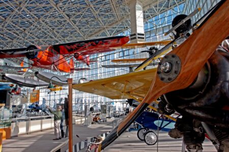 Aviation museum with various vintage aircraft displayed in a spacious, glass-ceilinged hall. The exhibit includes a bright orange plane and a wooden propeller in the foreground, with more airplanes and a visitor in the background.
