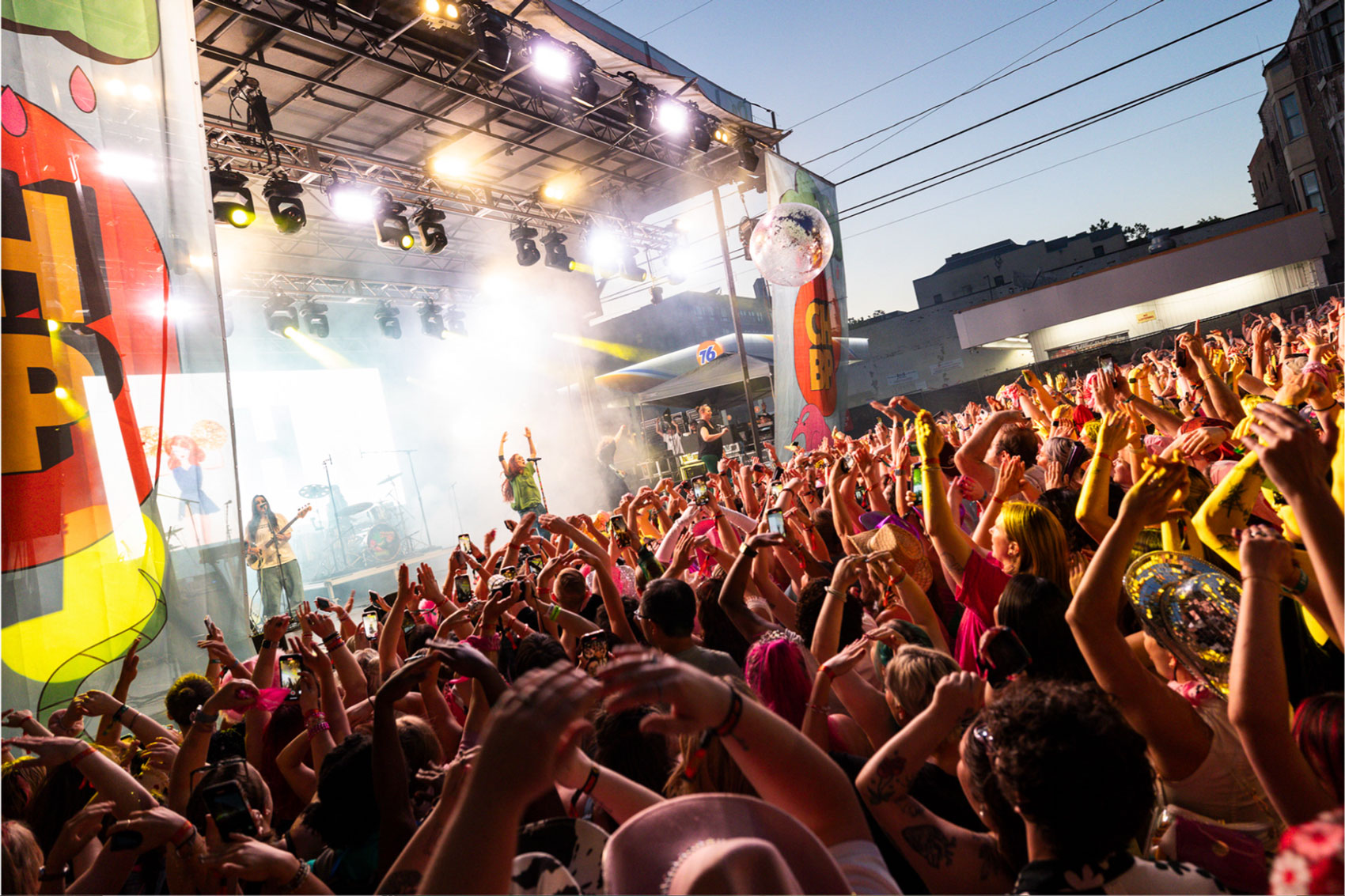 A vibrant outdoor concert scene with a lively crowd under colorful lighting. People raise their hands in excitement as performers are on stage. A large video screen and a disco ball add to the festive atmosphere.
