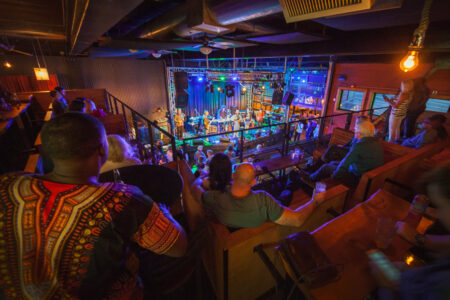 A vibrant indoor concert scene with a band performing on stage under colorful lights. The venue is filled with a diverse audience seated on wooden benches and standing, enjoying the live music. The atmosphere is lively and energetic.