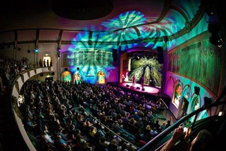 A vibrant, colorful theater scene with a full audience. Bright, multicolored lights create dynamic patterns on the domed ceiling. On stage, performers entertain under a dramatic, illuminated backdrop. The theater has ornate architectural details.