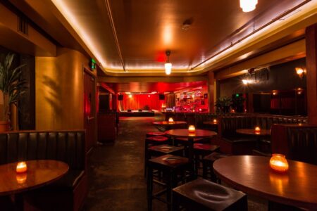 A cozy, dimly lit bar with small round tables and cushioned booths. Candles on tables create a warm ambiance. A stage with closed red curtains is at the far end, and the bar is illuminated by soft lighting.