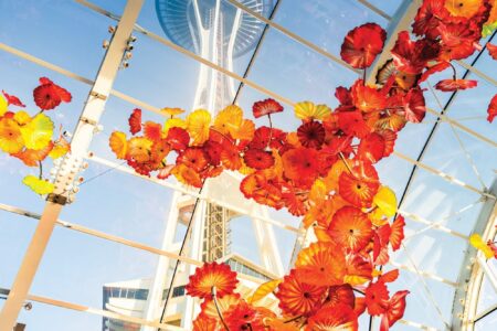 A view of the Space Needle seen through a glass ceiling adorned with vibrant red and yellow glass flowers, set against a clear blue sky.