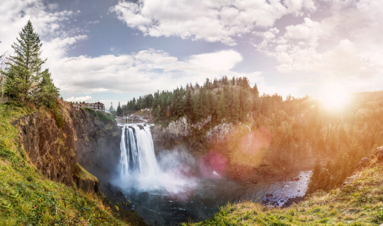 A majestic waterfall cascades over a cliff surrounded by lush forest under a partly cloudy sky. Sunlight shines brightly, creating a lens flare effect. A building is perched on the cliff's edge to the left.