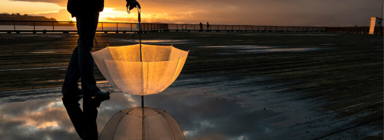 A person stands on a wet boardwalk at sunset, holding a transparent umbrella. The umbrella is open and its reflection is visible in a nearby puddle. Silhouetted figures stand in the distance against the orange sky.