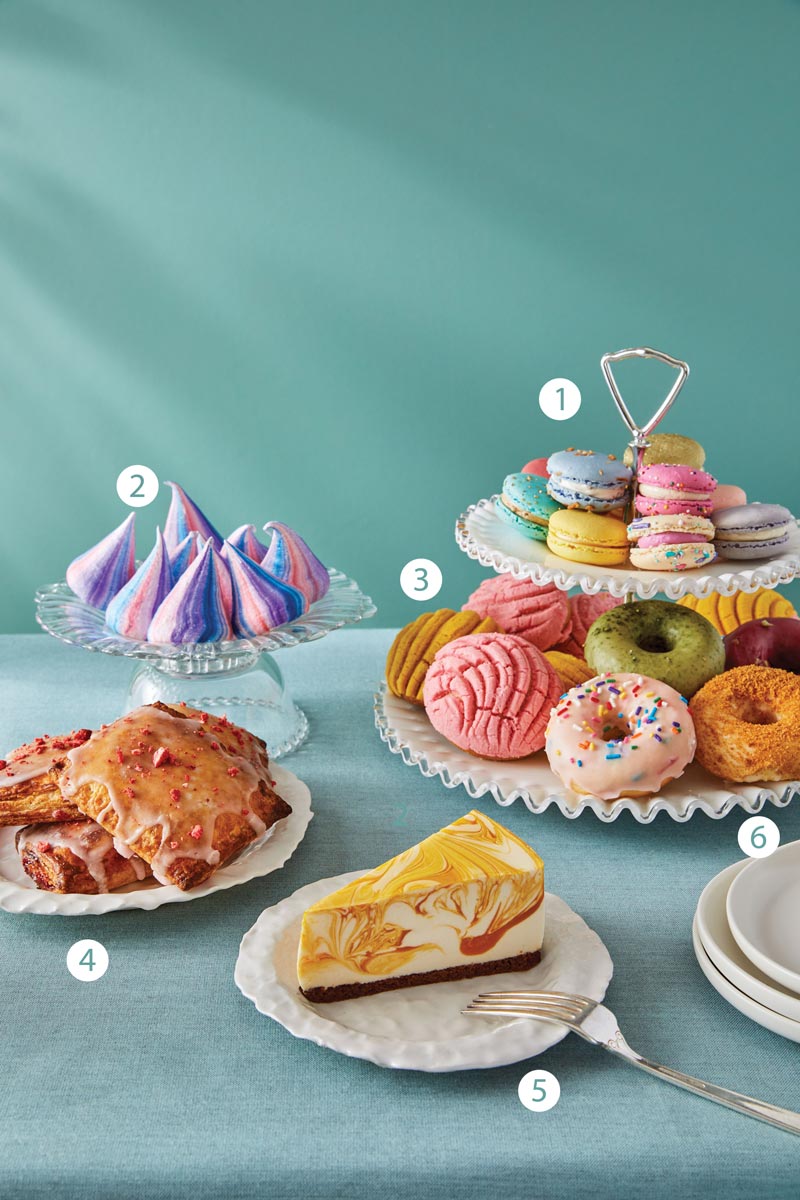 An arrangement of desserts on a table: a tiered stand with macarons and cookies, a plate of colorful swirl meringues, a dish with fried cookies, glazed donuts, croissants with toppings, and a slice of marbled cheesecake with a fork.