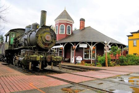 A vintage steam locomotive displayed in front of a historic red train station with a conical roof. The setting includes brick pathways, railroad tracks, and surrounding greenery under an overcast sky.