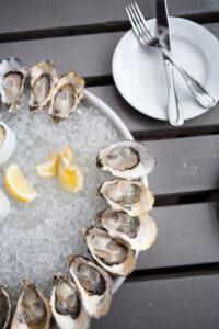 A round metal tray filled with ice, topped with a dozen opened oysters and lemon wedges. The tray is on a gray wooden table, with a white plate, fork, and knife nearby.