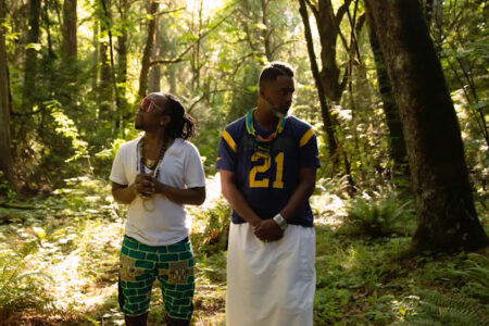 Two people stand in a sunlit forest. One wears a white shirt and turquoise pants with patterns, and the other wears a blue jersey with the number 21 and a white skirt. Both are gazing in different directions, surrounded by tall trees and greenery.