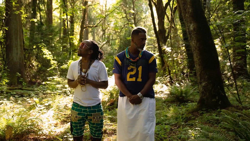 Two people stand in a sunlit forest. One wears a white shirt and turquoise pants with patterns, and the other wears a blue jersey with the number 21 and a white skirt. Both are gazing in different directions, surrounded by tall trees and greenery.