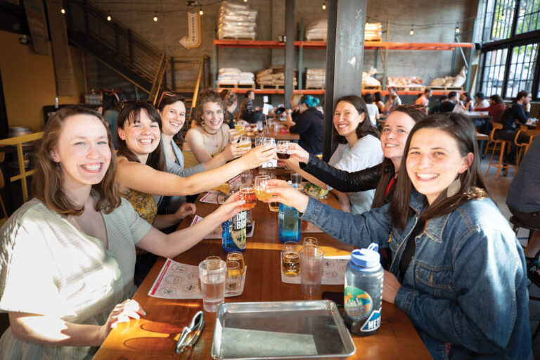 A group of people sitting at a long wooden table in a bustling restaurant, smiling and raising their glasses for a toast. The room has industrial decor with shelves stocked with bags. Sunlight streams through large windows.