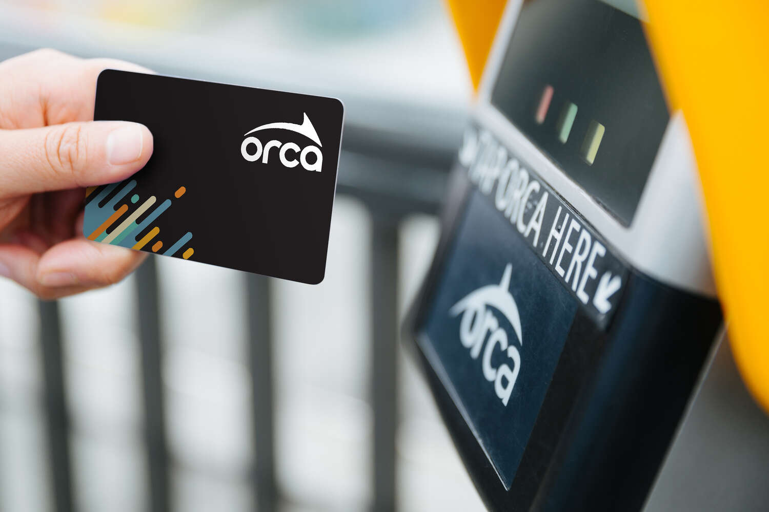 A person holds an ORCA card near a card reader, preparing to tap for transit fare payment. The card features a logo with a stylized orca and colorful lines.