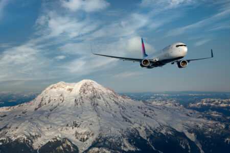 A commercial airplane flies past a snow-capped mountain under a clear blue sky, with scattered clouds enhancing the scenic view. The majestic landscape features rugged, snowy peaks and rocky terrain.