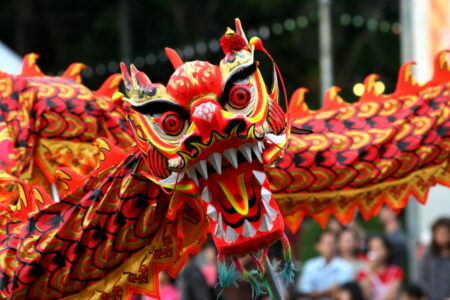 A vibrant, colorful dragon puppet with large eyes and sharp teeth, featuring intricate red, orange, and yellow scales, is being paraded outdoors. People in the blurred background are watching the performance.