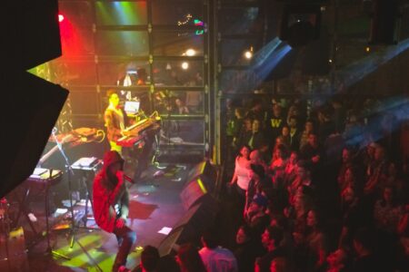 A lively indoor concert with colorful stage lights. A performer sings energetically into a microphone while playing keyboards on stage. Below, a large crowd watches and enjoys the music.