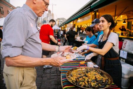 A woman serves food from a large pan of paella to an older man at an outdoor market. The scene is lively with people in the background, and colorful tablecloths and lights decorate the stalls.