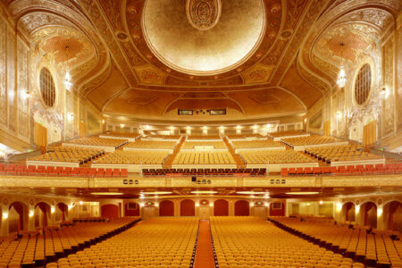 Ornate theater interior with rows of plush, golden seats on multiple levels. Intricate designs adorn the ceiling and walls. Warm lighting enhances the opulent atmosphere. A large stage is visible at the front.