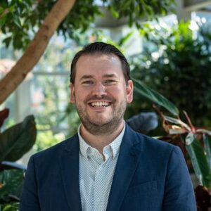 A person with a beard is smiling, wearing a blue blazer and white shirt, standing in front of lush green plants in a bright location.
