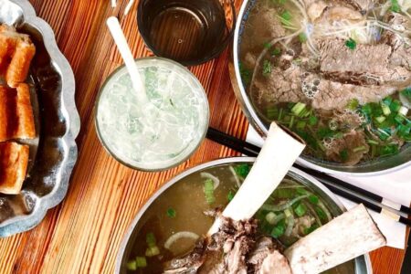 Two bowls of beef pho with bone-in meat and herbs on a wooden table, accompanied by a glass of iced drink, a plate of crispy rolls, chopsticks, and a small dish of fresh vegetables.