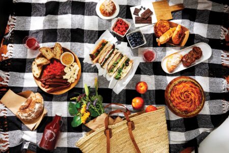 A picnic spread on a checkered blanket featuring sandwiches, bread, fruits, chocolate, and pastries. A woven basket, flowers, and a bottle are part of the arrangement. The scene conveys a cozy and inviting outdoor meal setting.