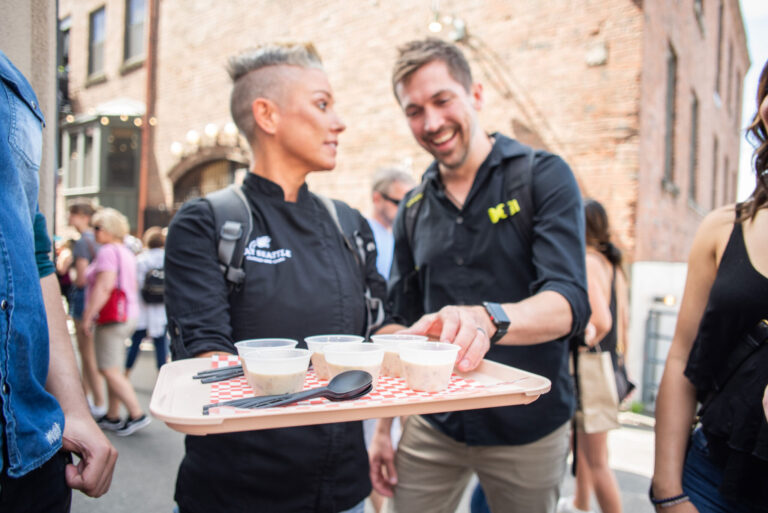 Two people outside, one holding a tray with small cups and a spoon. They appear to be smiling and chatting about exciting things to do in Seattle. The scene is lively, with a brick wall and other people in the background.