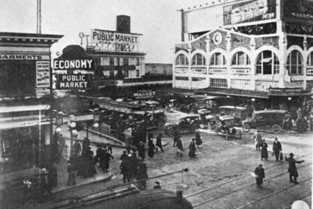 Black and white image of a bustling public market with vintage cars parked and people walking. Prominent signs read 