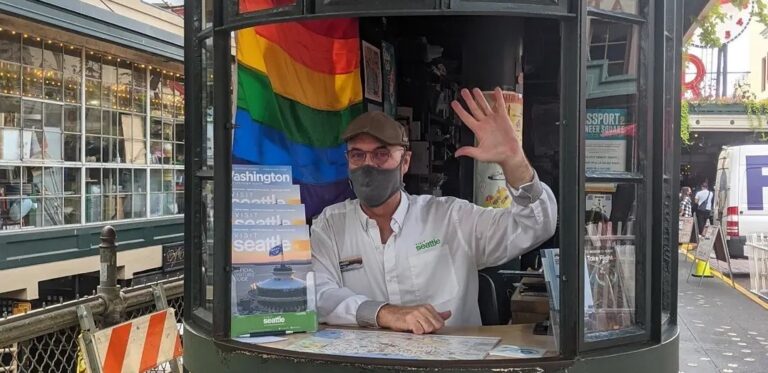 A person wearing a cap and mask waves from inside a small circular kiosk. A rainbow flag is displayed behind them. Maps and brochures about Seattle are visible on the desk. The kiosk is surrounded by bustling activity and festive lights.