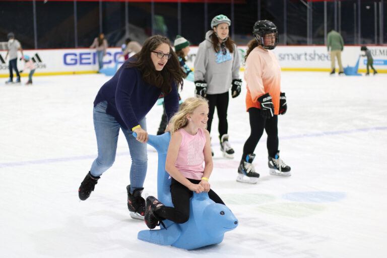 A photo of people skating on the ice at the Kraken Community Iceplex. A woman pushes a young girl on a blue seal seat across the ice.
