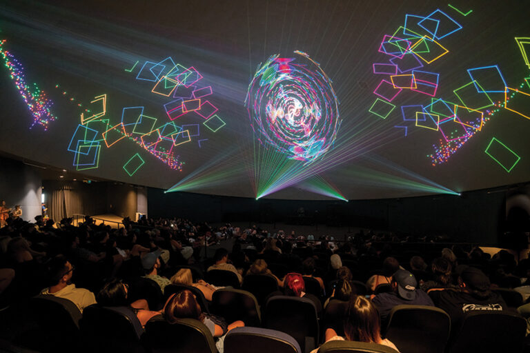 A photo of a laser show at the Pacific Science Center's Laser Dome. The audience sits in a dark room while rainbow colored lights project onto the walls and ceiling.