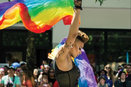A person in a sheer black outfit waves a large rainbow flag at an outdoor event. A crowd watches in the background, some holding smaller rainbow flags. Trees and a building are visible behind the crowd.