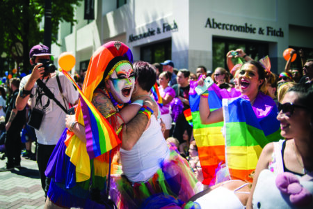 A lively group celebrates at a pride parade. Two people hug, wearing colorful outfits and rainbow flags. Joyful onlookers cheer and capture the moment with phones. A store is visible in the background.